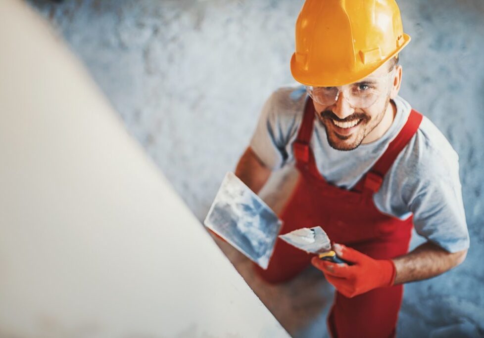 Closeup top view of a construction worker finishing a facade in an interior drywall. He's smiling and looking at camera. Wearing red uniform and yellow work helmet.